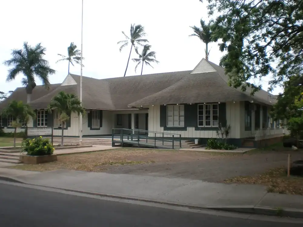 a single-story wooden plantation-era school building with a wide hip roof and palm trees in the historic Ewa plantation village