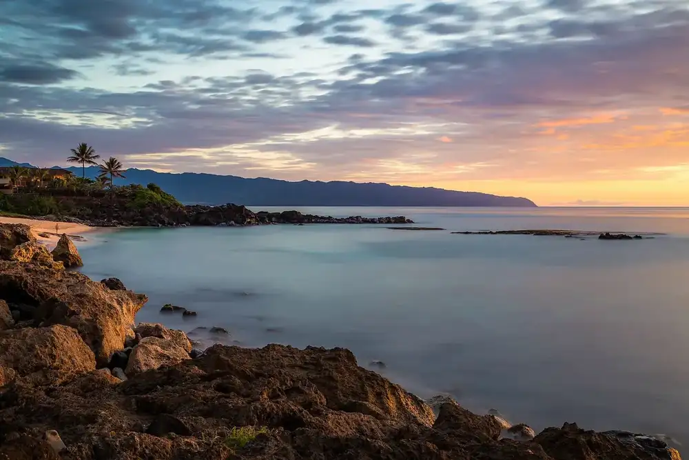 a rocky shoreline and calm bay at Haleiwa on the North Shore at sunset, with mountains along the coast