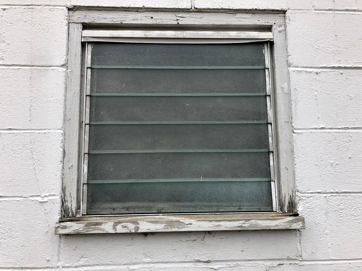 a jalousie window with horizontal louvered glass slats in a worn white wooden frame set in a painted block wall