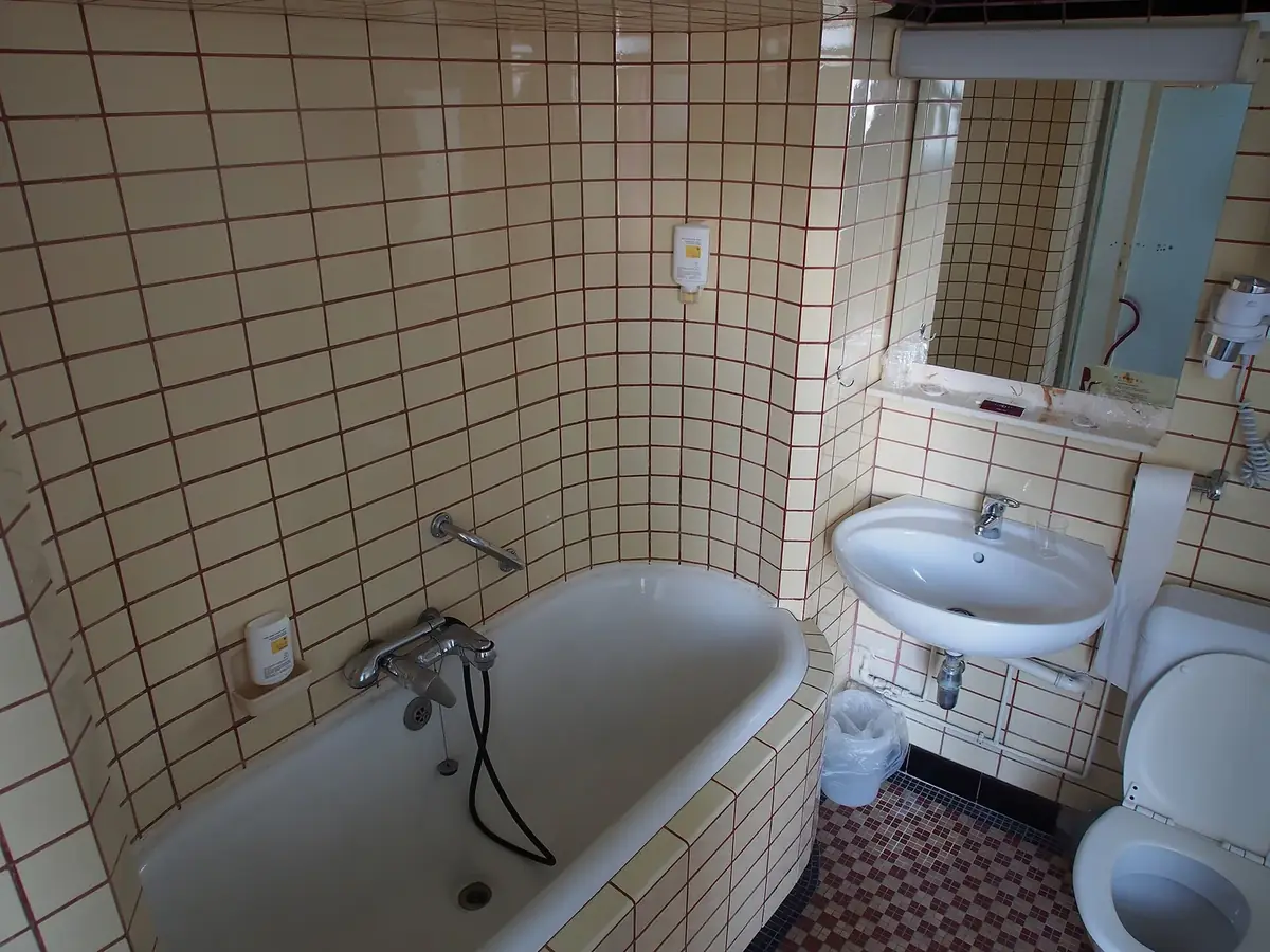 an older bathroom tiled floor to ceiling in cream square tiles with dark grout, with a tub, a wall sink and a toilet