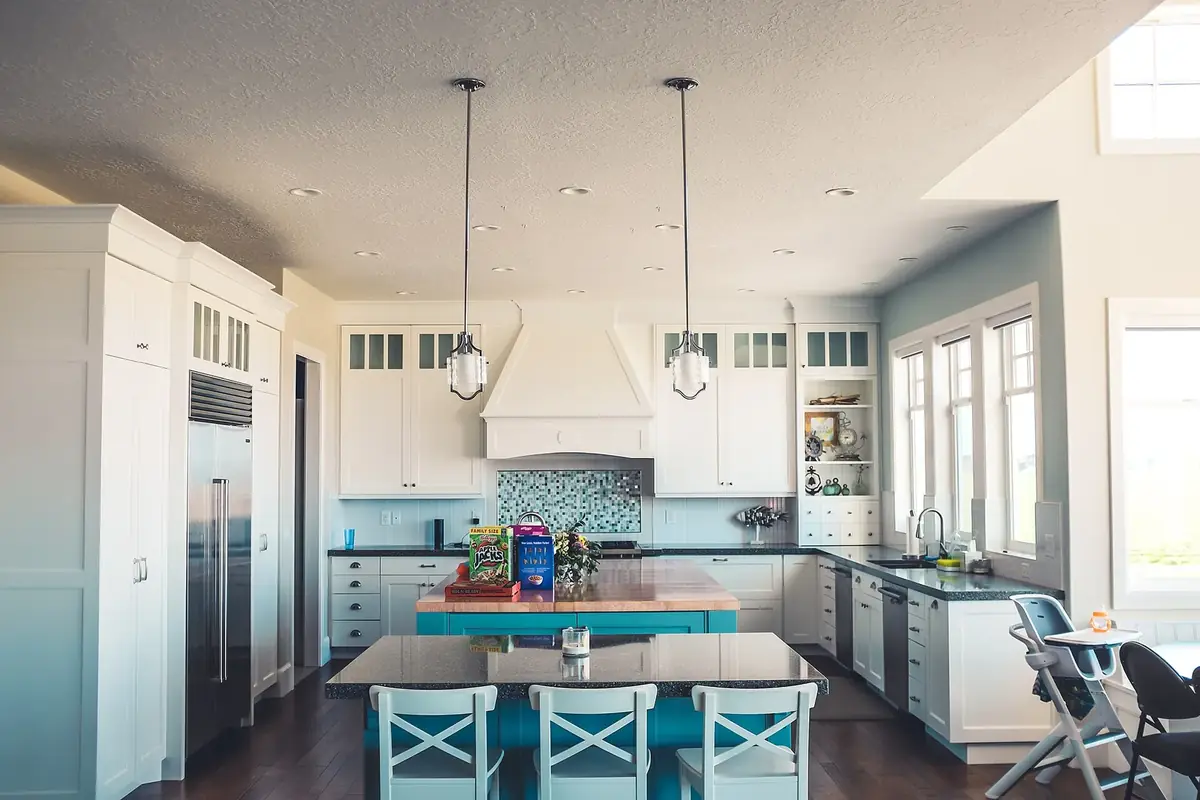 a bright kitchen with white cabinets to the ceiling, a range hood, pendant lights and a teal island with stools