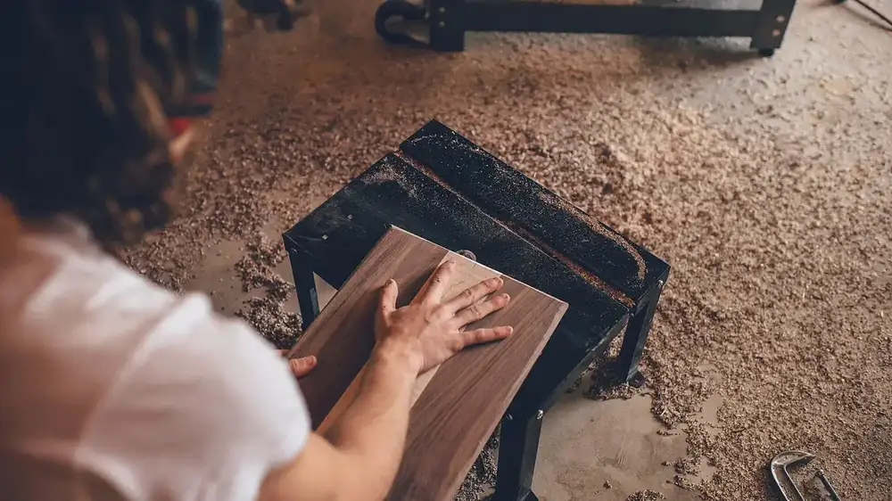 over-the-shoulder view of a woodworker pressing a walnut board flat on a small black work stand, on a sawdust-covered workshop floor with a C-clamp nearby